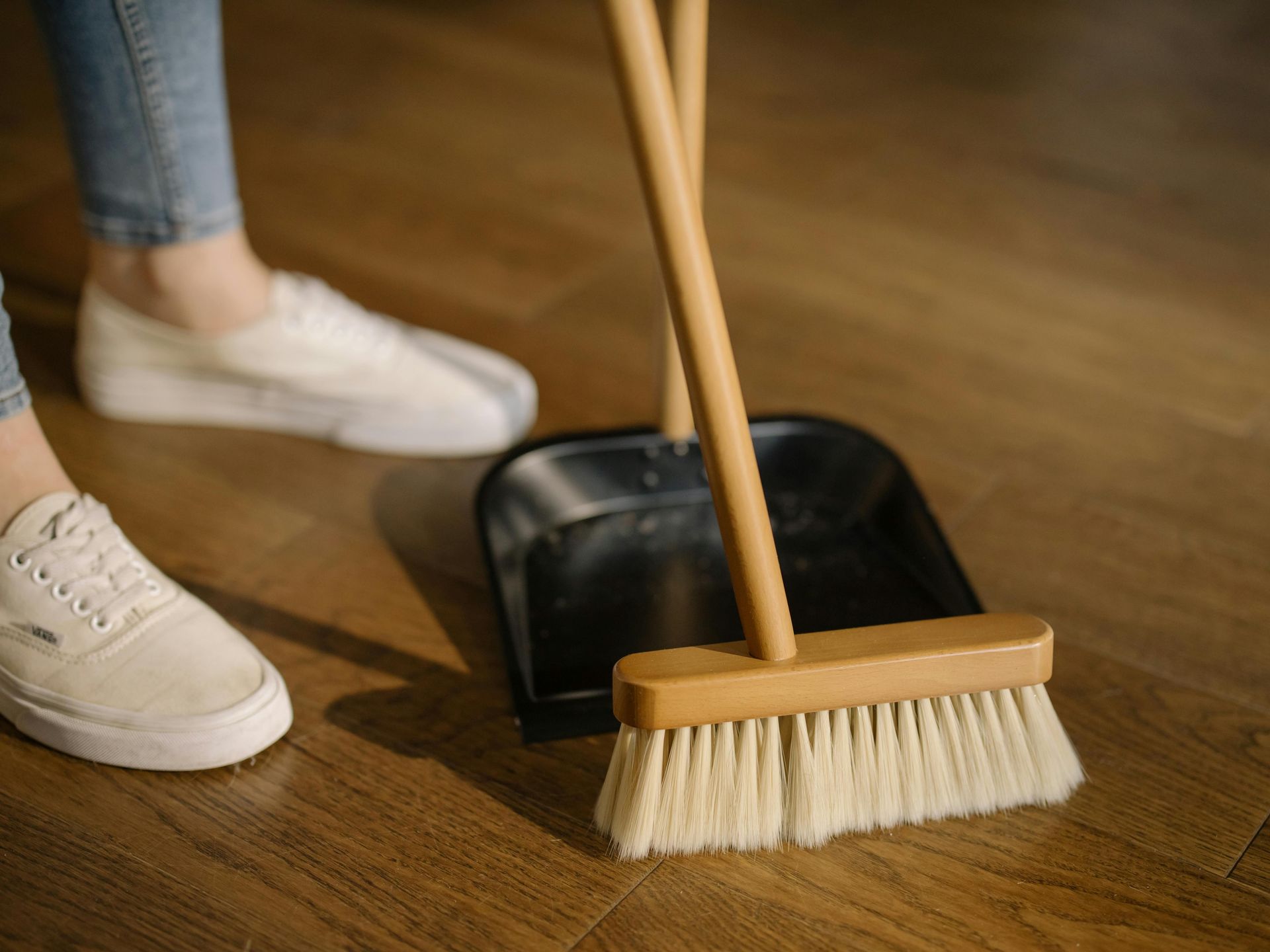 Person sweeping wooden floor with a broom and dustpan. White sneakers and jeans visible.